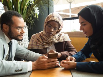 Excited diverse coworkers watching content on smartphone. Businessman and Muslim businesswomen sitting in cafe, man showing phone screen to colleagues. Digital technology concept